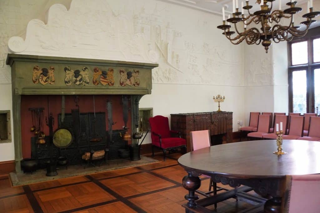 Der prunkvolle Rittersaal im Soest Burghofmuseum zeigt einen großen, verzierten Kamin mit Wappen, einen massiven runden Holztisch und historische Möbel unter einer stuckverzierten Wand. Ein prachtvoller Kronleuchter hängt von der Decke über dem Parkettboden des historischen Raumes.