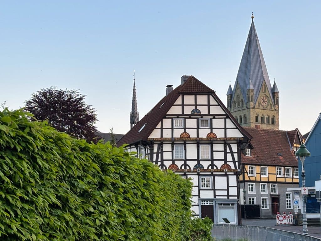Blick über eine grüne Hecke auf ein historisches Fachwerkhaus in Soest, hinter dem die markante Turmspitze des St.-Patrokli-Doms in den klaren Abendhimmel ragt.