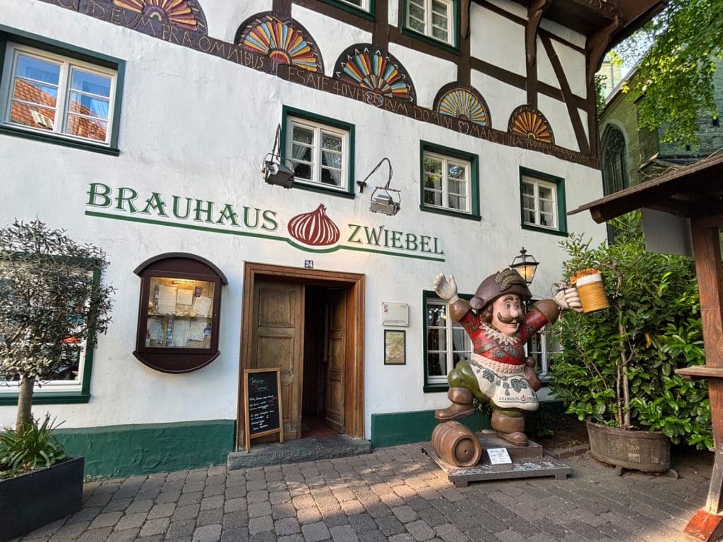 Eingang des Brauhaus Zwiebel in Soest mit einer weißen Fachwerkfassade, grünen Fensterrahmen und einer großen, fröhlichen Statue eines Mannes mit Bierkrug vor der Tür. Über dem Eingang prangt der Schriftzug „Brauhaus Zwiebel“ mit einem roten Zwiebel-Logo.