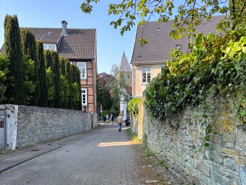 Blick durch eine gepflasterte Gasse in Soest während einer Foodtour, gesäumt von historischen Steinmauern und Fachwerkhäusern, mit Passanten und einem Kirchturm im Hintergrund.