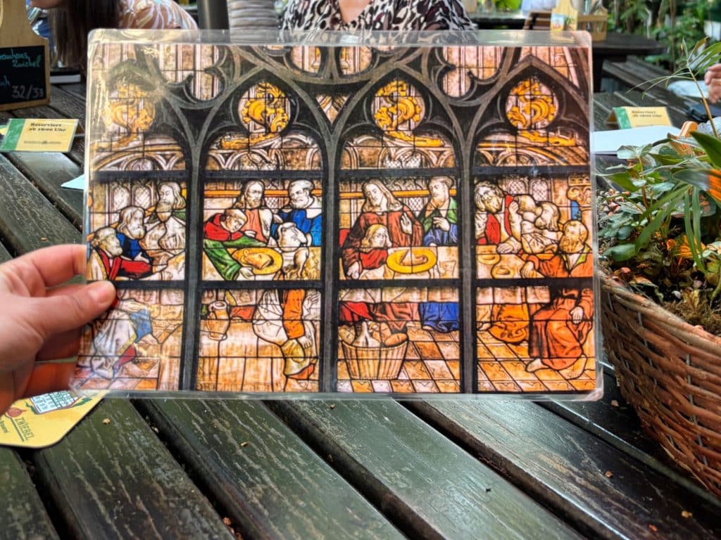 Eine Person hält eine laminierte Abbildung des „Westfälischen Abendmahls“ aus Soest über einen rustikalen Holztisch, auf der Jesus und seine Jünger bei einer Mahlzeit mit regionalen Spezialitäten wie Schweinskopf und Pumpernickel anstelle von Brot und Wein dargestellt sind.