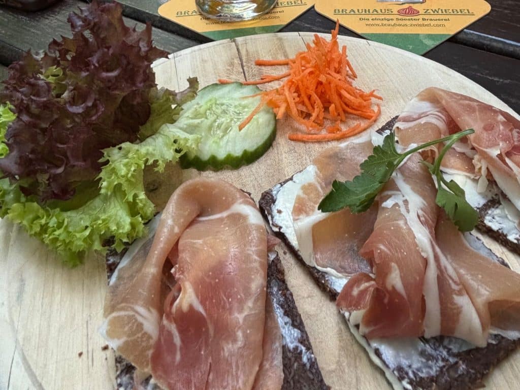 Vorspeise der Menüsafari in Soest im Brauhaus Zwiebel: Pumpernickel mit weißem Aufstrich und Schinken, garniert mit Salat, Gurke und geraspelten Karotten auf einem Holzbrett.