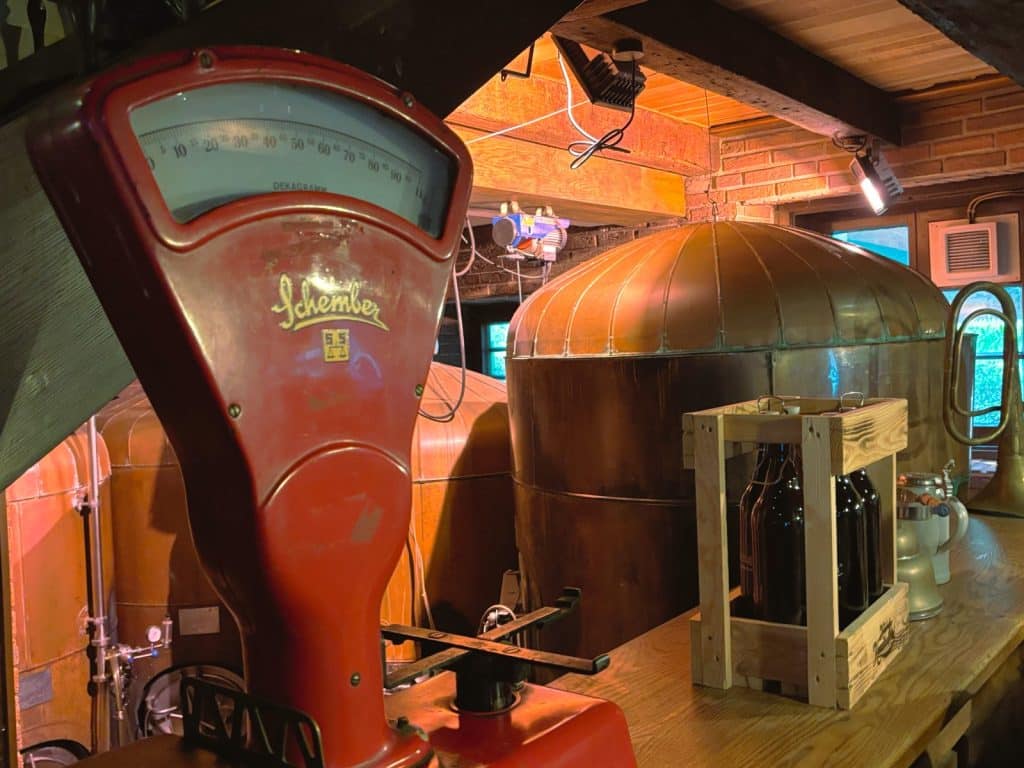 Eine rote, antike Schember-Waage steht im Vordergrund einer Brauerei in Soest, in deren Hintergrund große kupferne Braukessel und eine Holzkiste mit Bierflaschen zu sehen sind.