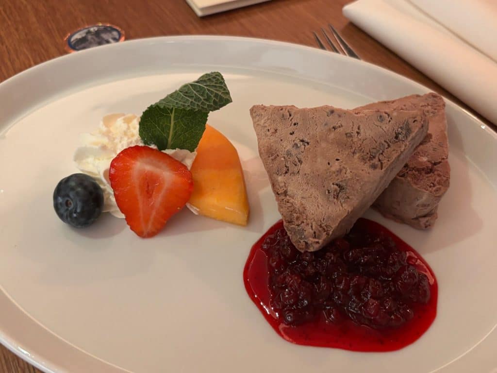 Ein Dessertteller im Pilgrimhaus in Soest mit zwei dreieckigen Stücken Pumpernickelparfait auf einem roten Beerenkompott, garniert mit einer Erdbeerscheibe, einer Blaubeere, einem Pfirsichspalt, Sahne und einem Minzblatt.