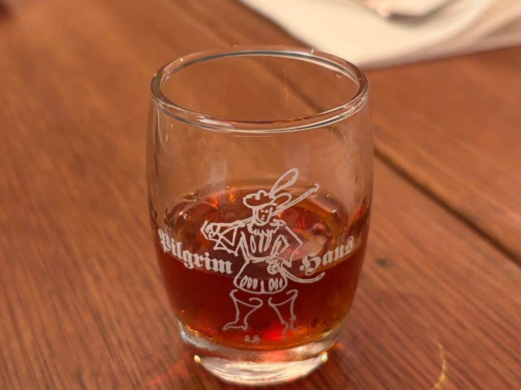 Ein kleines Schnapsglas mit einer bernsteinfarbenen Flüssigkeit steht auf einem Holztisch, verziert mit dem weißen Logo des Pilgrimhauses in Soest, das eine historische Figur zeigt.