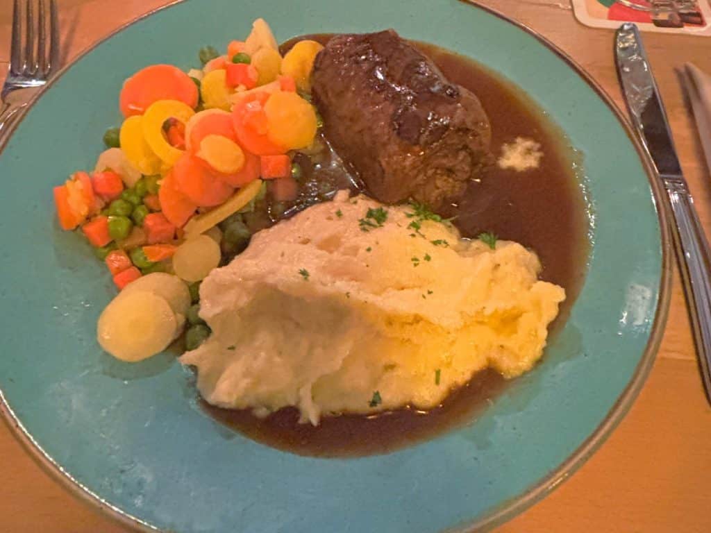 Ein Hauptgericht der Menüsafari in Soest im Restaurant Wilder Mann, bestehend aus einer Rinderroulade in dunkler Soße, serviert mit cremigem Kartoffelstampf und buntem Mischgemüse auf einem türkisfarbenen Teller.