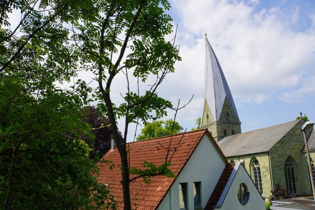 Blick über grüne Bäume und ein Haus mit rotem Ziegeldach auf die St.-Andreas-Kirche in Soest mit ihrem markanten schiefen Turmhelm unter leicht bewölktem Himmel.