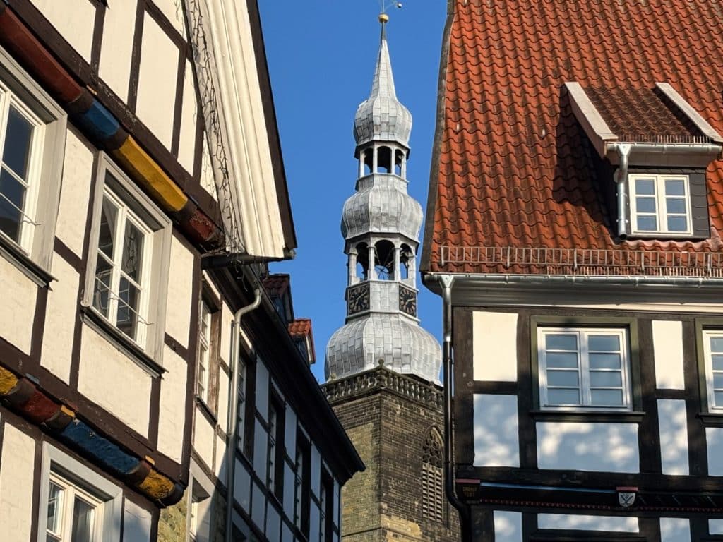 Blick durch eine schmale Gasse mit historischen Fachwerkhäusern auf den markanten, mehrstufigen Turmhelm der St. Petri-Kirche in Soest unter strahlend blauem Himmel.
