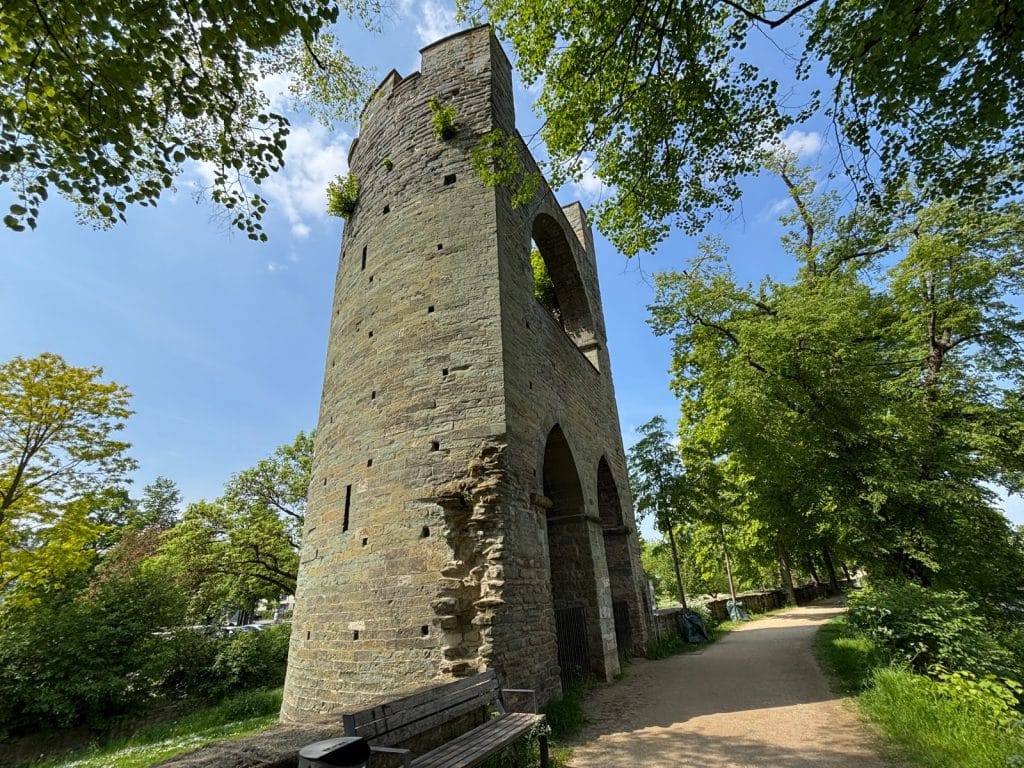 Der mittelalterliche Kattenturm an den Wallanlagen Soest ragt als steinerner Wehrturm mit Rundbogenöffnungen hinter einer Parkbank an einem von Bäumen gesäumten Spazierweg empor.