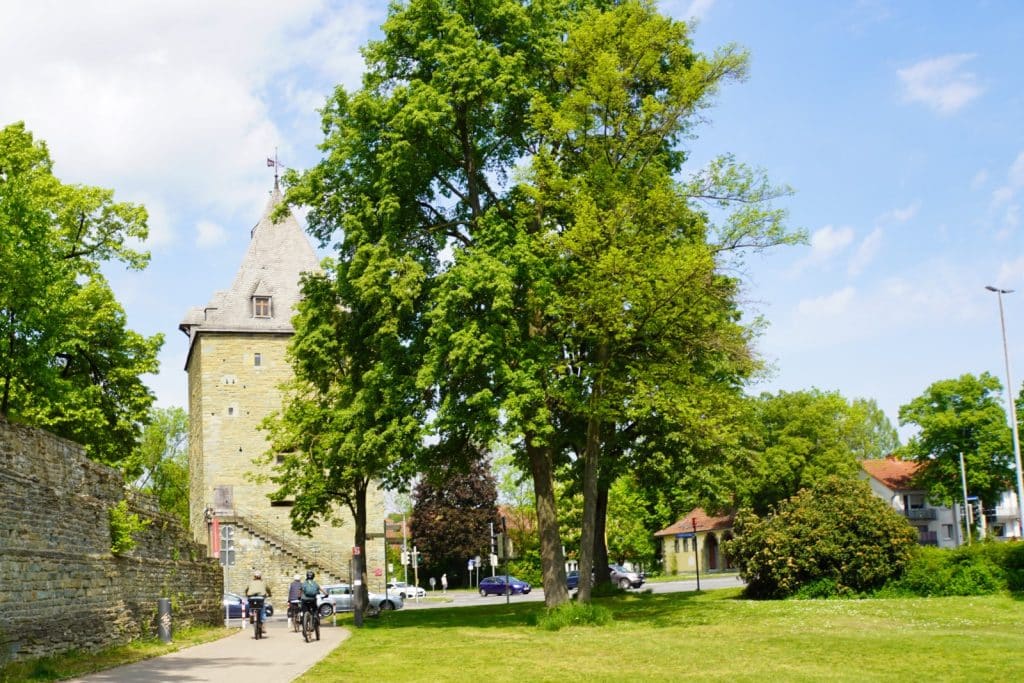 Wallanlagen Soest : Ein gepflasterter Weg führt an der historischen Stadtmauer und dem steinernen Osthofentor am Soester Wall vorbei, während Radfahrer unter hohen grünen Bäumen in Richtung einer Straße fahren.
