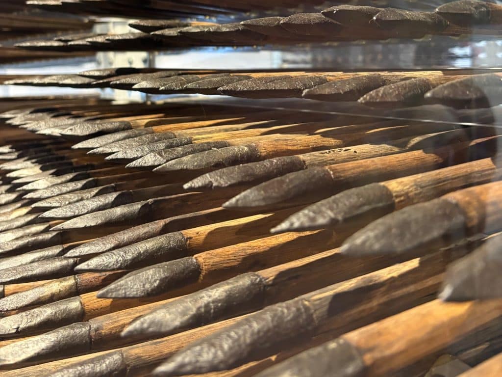 Zahlreiche historische Armbrustpfeile mit dunklen Metallspitzen und hölzernen Schäften sind in einer Vitrine im Soester Stadttor dicht an dicht aufgereiht.