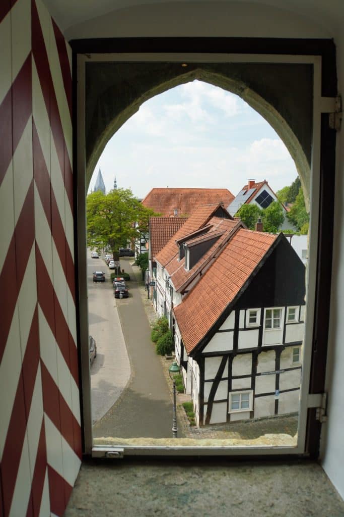 Blick aus einem bogenförmigen Fenster des Soester Wall-Stadttores auf eine Straße mit historischen Fachwerkhäusern und geparkten Autos unter einem leicht bewölkten Himmel. Links im Vordergrund ist ein rot-weiß gestreifter Fensterladen zu sehen, der den Blick auf die Altstadt einrahmt.