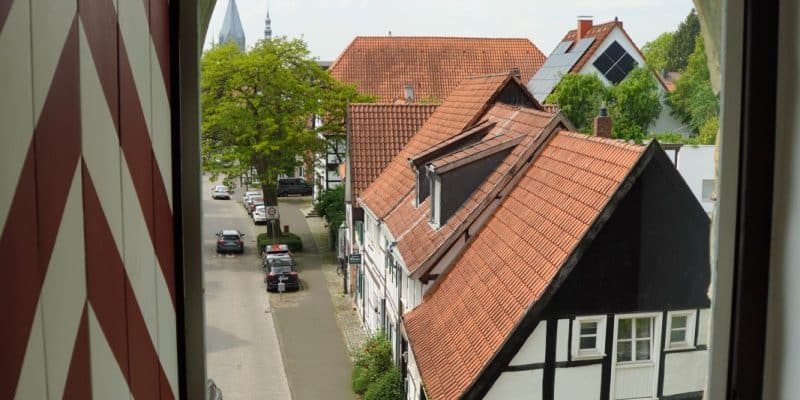 Blick aus einem bogenförmigen Fenster des Soester Wall-Stadttores auf eine Straße mit historischen Fachwerkhäusern und geparkten Autos unter einem leicht bewölkten Himmel. Links im Vordergrund ist ein rot-weiß gestreifter Fensterladen zu sehen, der den Blick auf die Altstadt einrahmt.