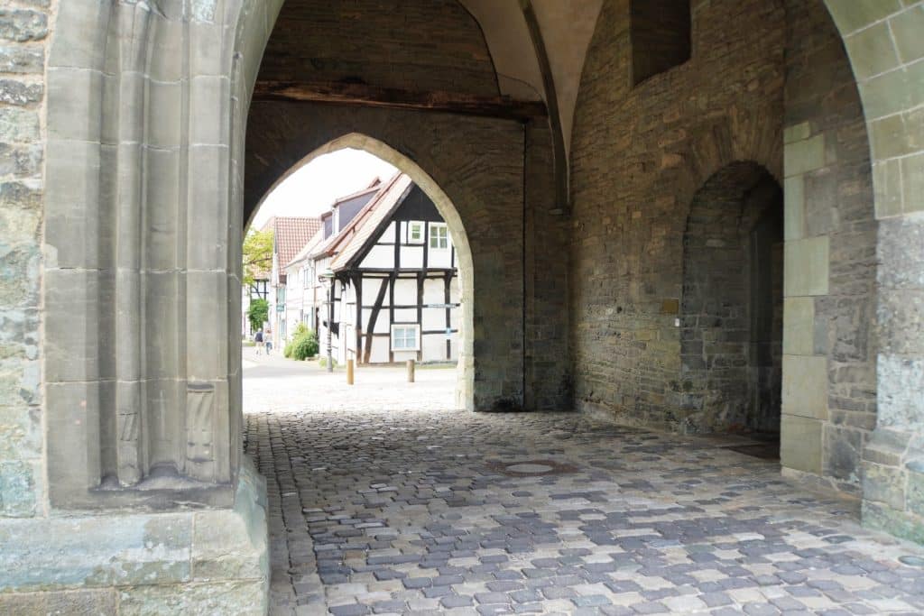 Blick durch ein steinernes Stadttor an der Soester Wallstraße auf eine gepflasterte Gasse mit einem historischen Fachwerkhaus im Hintergrund.