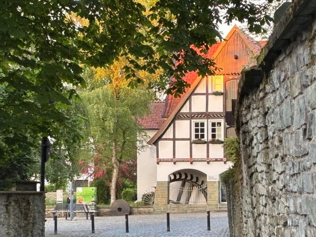 Ein historisches Fachwerkhaus in Soest mit einem großen hölzernen Mühlrad hinter einem Torbogen, eingerahmt von grünen Bäumen und einer alten Steinmauer im Vordergrund.