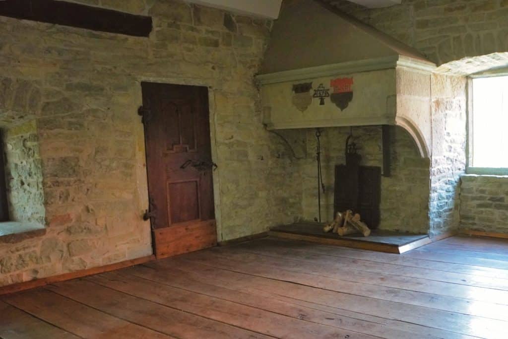 Innenansicht des romanischen Hauses in Soest mit freigelegten Steinwänden, einem breiten Holzdielenboden und einem großen Kamin mit Wappenverzierung neben einer rustikalen Holztür.