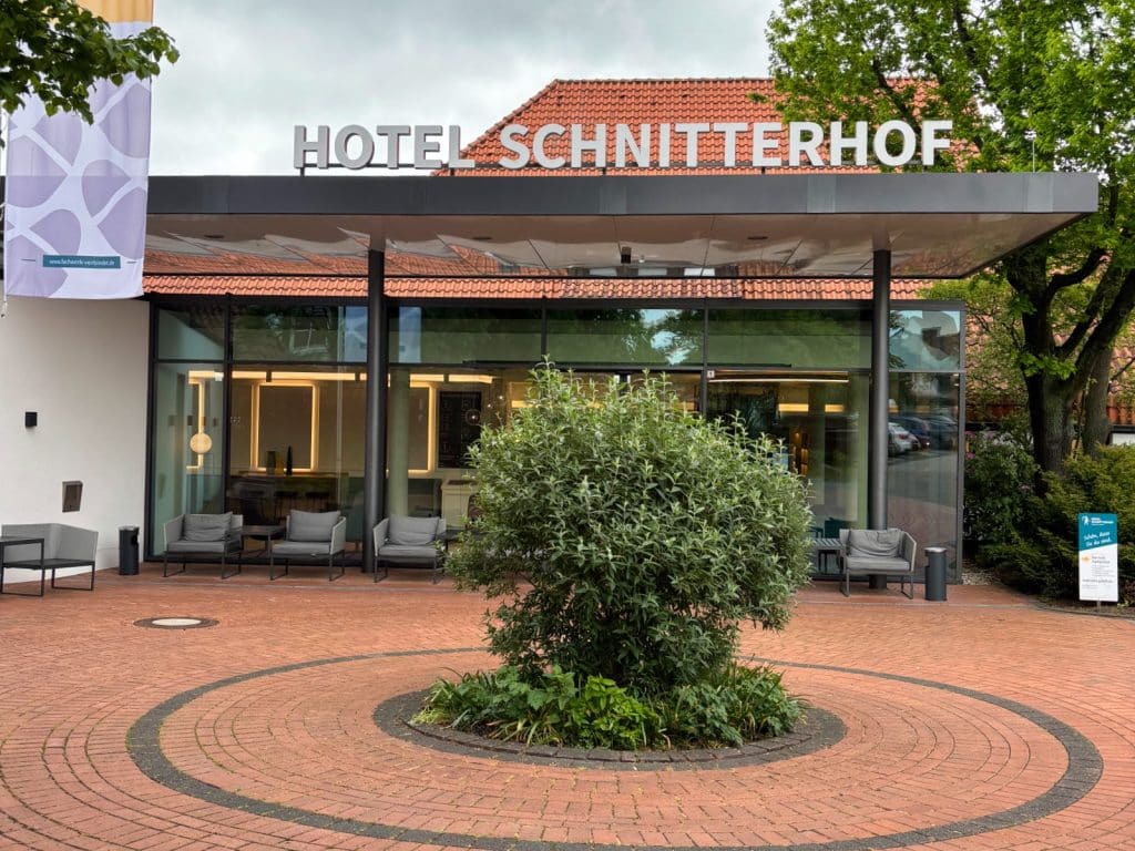 Der moderne Eingangsbereich des Hotels Schnitterhof in Bad Sassendorf zeigt eine Glasfront unter einem Vordach mit dem Schriftzug des Hotels, davor befindet sich ein gepflasterter Vorplatz mit einer runden Grünanlage im Zentrum.