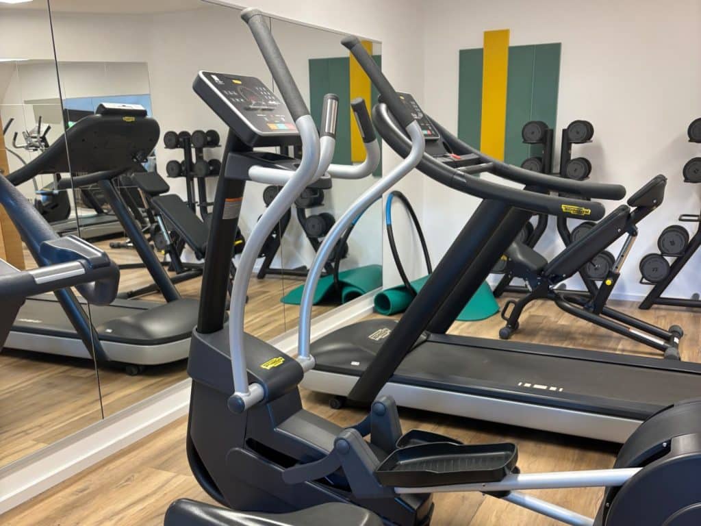 Der moderne Fitnessraum im Hotel Schnitterhof in Bad Sassendorf ist mit hochwertigen Technogym-Geräten ausgestattet, darunter ein Crosstrainer im Vordergrund, Laufbänder und Hantelstationen vor einer verspiegelten Wand.