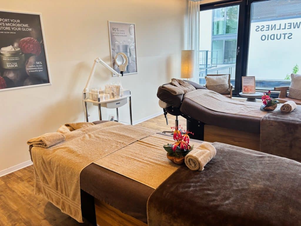 Ein heller Behandlungsraum für Massagen in Hotel Schnitterhof mit zwei parallel angeordneten, gemütlich hergerichteten Massageliegen vor einem großen Fenster. Ein Rollwagen mit Pflegeprodukten, eine Lupenleuchte und Werbeplakate für Kosmetikbehandlungen ergänzen das einladende Wellness-Ambiente.
