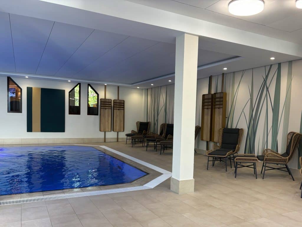 Ein moderner Innenpool im Hotel Schnitterhof mit blau leuchtendem Wasser, umgeben von einer hellen Terrasse mit komfortablen Lounge-Sesseln vor einer Wand mit Gräser-Motiv.
