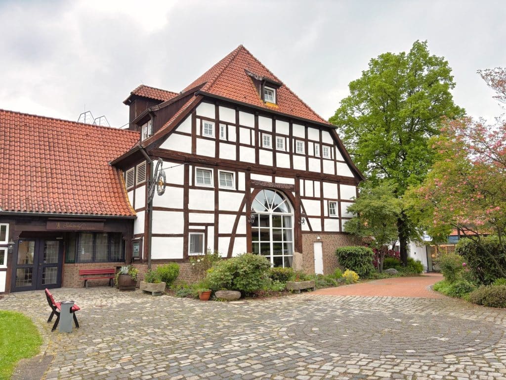 Außenansicht eines historischen Fachwerkhauses in Bad Sassendorf, das als Hotel und Restaurant genutzt wird, mit einem gepflasterten Vorplatz, roten Ziegeldächern und einer großen Rundbogen-Glasfront.