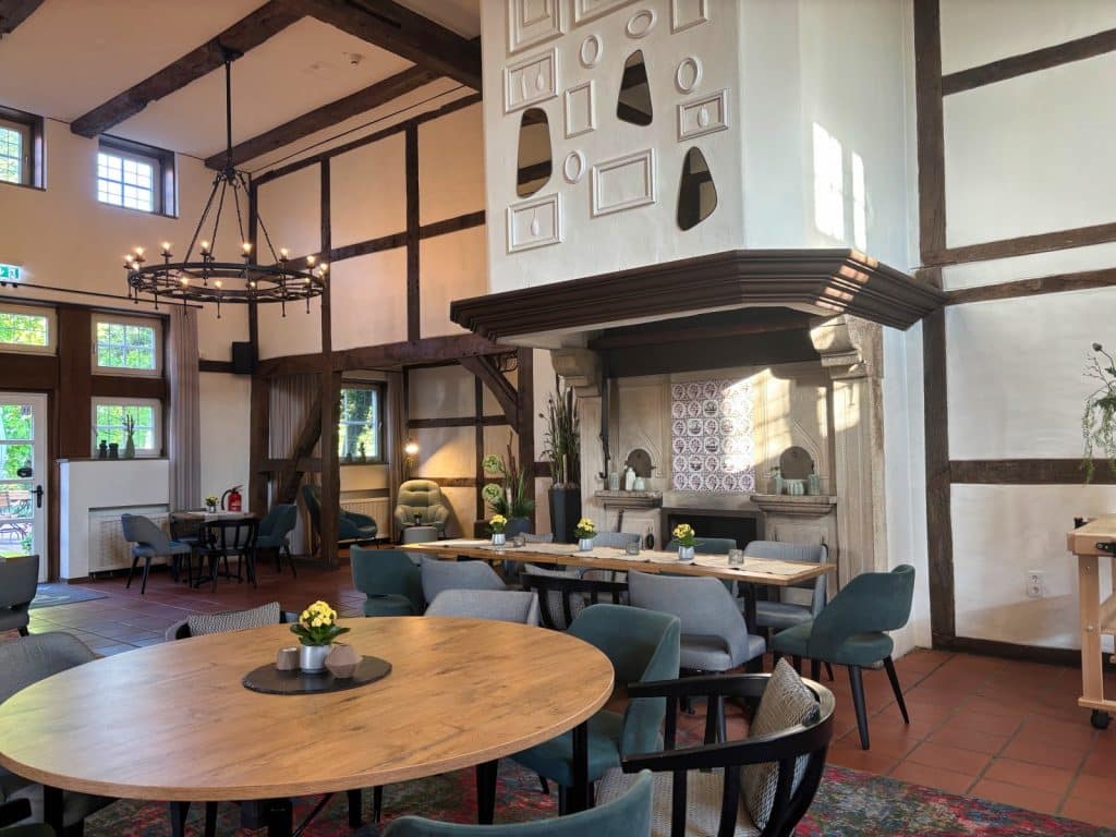 Ein gemütlicher Saal in Bad Sassendorf mit rustikalem Fachwerk, einem großen Kamin mit dekorativen Wandelementen und verschiedenen runden sowie eckigen Holztischen mit modernen blauen Polsterstühlen. Ein großer schmiedeeiserner Kronleuchter hängt von der hohen Decke mit dunklen Holzbalken herab und beleuchtet den einladenden Gastraum.