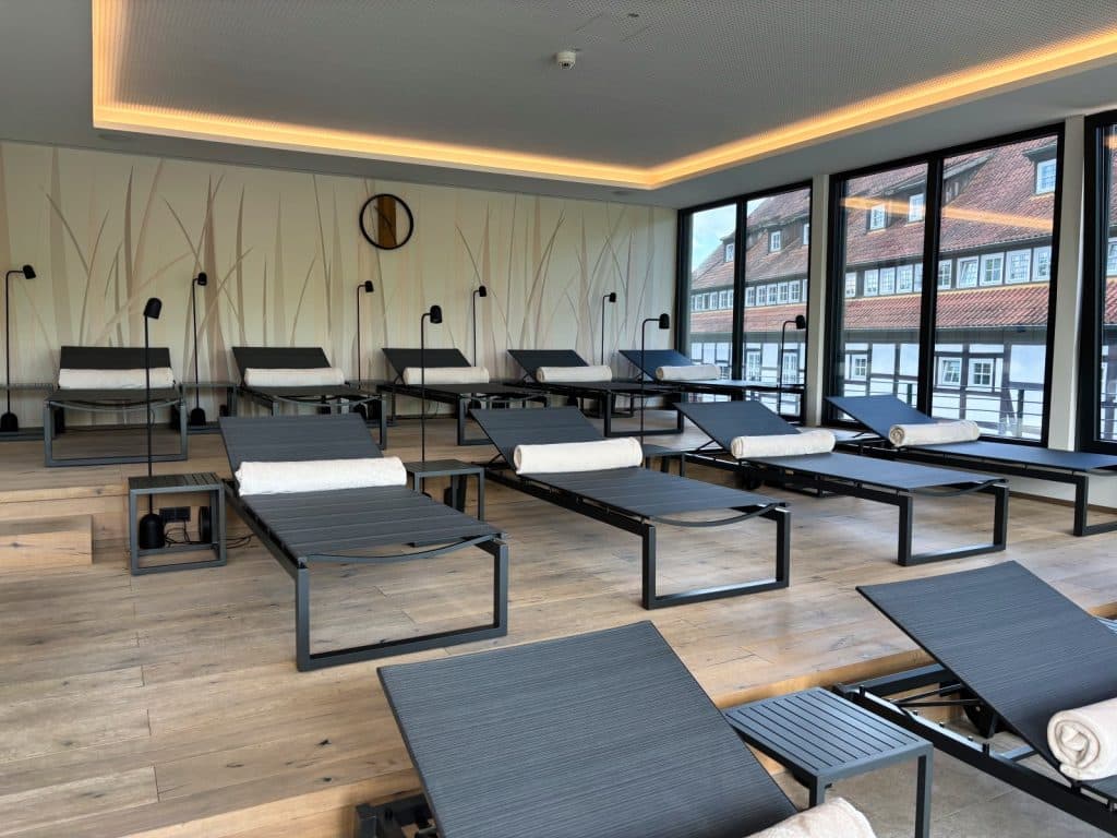 Ein moderner Ruheraum im Hotel in Bad Sassendorf mit mehreren dunklen Liegen, weißen Nackenrollen und Blick durch große Fenster auf historische Fachwerkhäuser. Die Wände sind mit einem dezenten Gräser-Motiv gestaltet und eine indirekte Deckenbeleuchtung sorgt für eine entspannte Atmosphäre.