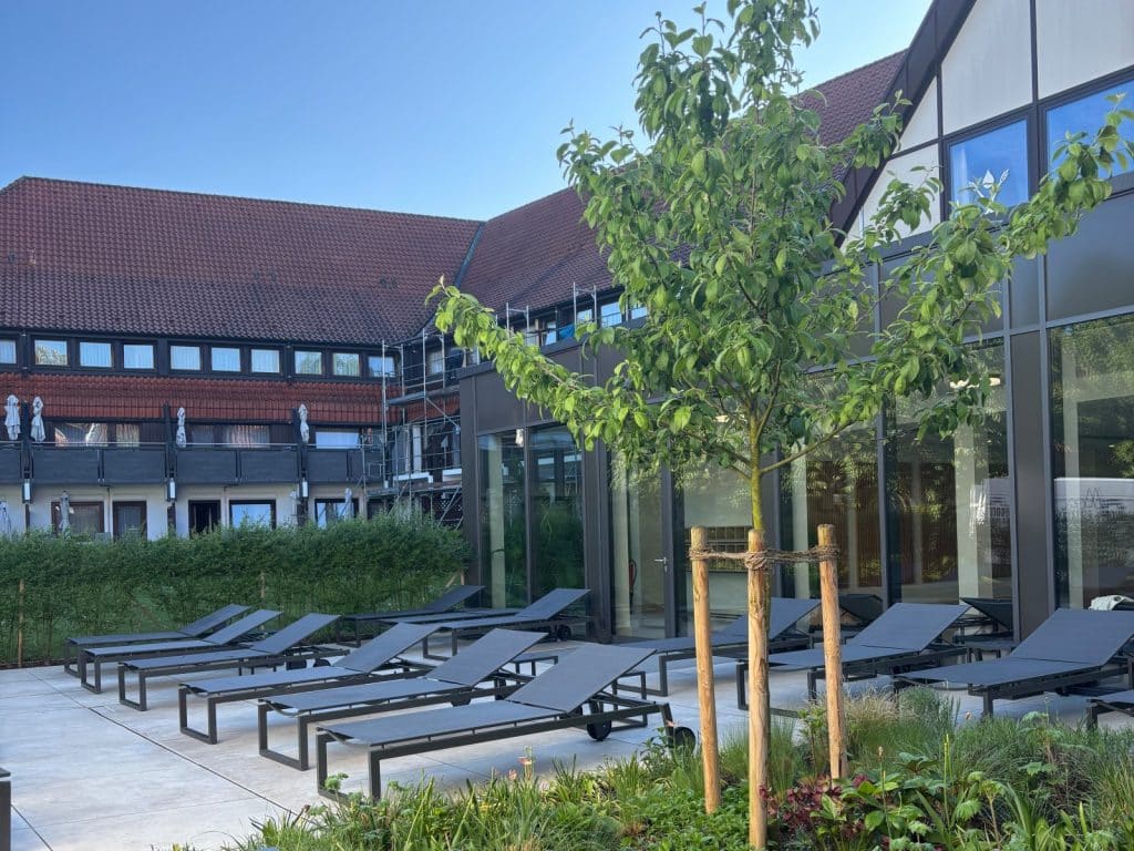 Die moderne Sonnenterrasse in Bad Sassendorf ist mit zahlreichen dunklen Liegestühlen ausgestattet und wird von einem jungen Baum sowie einer gepflegten Gartenanlage gesäumt.