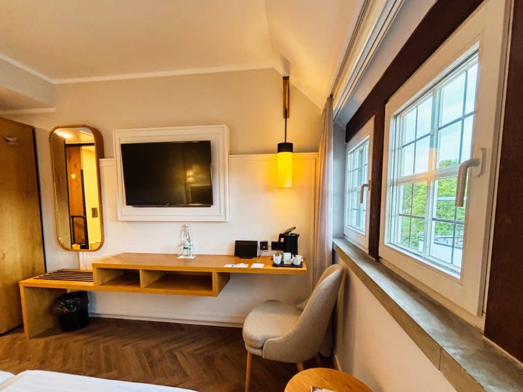 Ein modern eingerichtetes Hotelzimmer in Bad Sassendorf mit einem wandmontierten Fernseher, einem hellen Holzschreibtisch, einem bequemen Sessel und einem Fenster mit Blick ins Grüne.