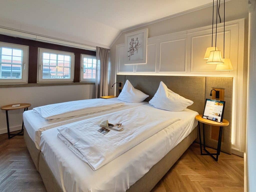 Ein modern eingerichtetes Hotelzimmer im Schnitterhof mit zwei zusammengeschobenen Einzelbetten, weißer Bettwäsche, indirekter Beleuchtung am gepolsterten Kopfteil und drei hängenden Designerlampen über dem Nachttisch.