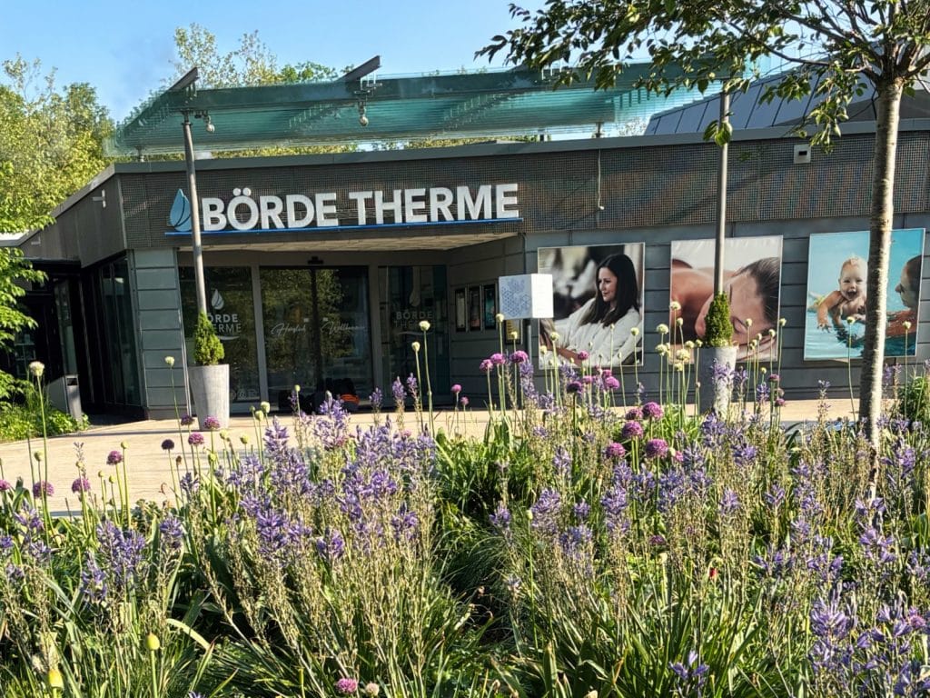 Außenansicht des modernen Eingangsgebäudes der Börde Therme Bad Sassendorf, davor ein blühendes Beet mit violetten Blumen.