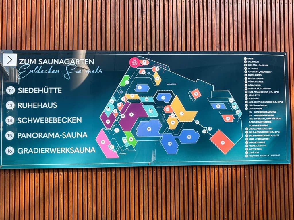 Ein Orientierungsplan des Saunagartens der Börde Therme Bad Sassendorf auf einer Glastafel vor einer Holzfassade zeigt eine farbige Übersichtskarte der Anlage mit nummerierten Stationen und einer Legende. Links sind ausgewählte Ziele wie die Siedehütte, das Ruhehaus und die Panorama-Sauna groß hervorgehoben.