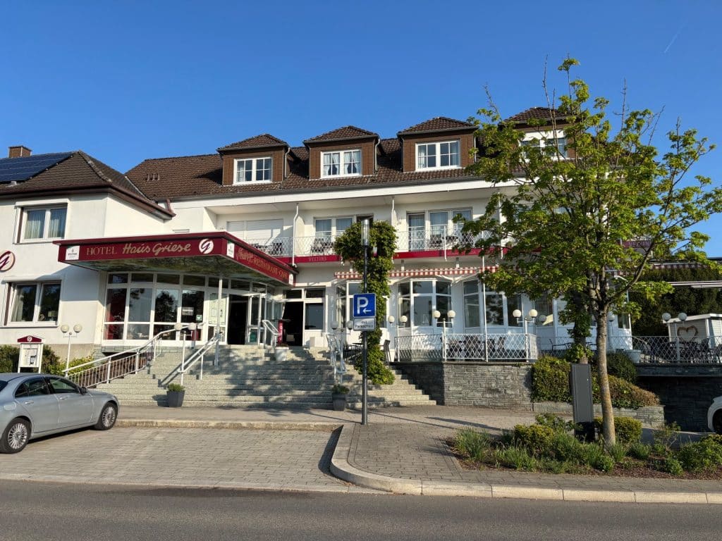 Das Hotel Haus Griese am Möhnesee präsentiert sich als weißes, mehrstöckiges Gebäude mit einer breiten Treppe zum Eingangsbereich und einer Außenterrasse unter strahlend blauem Himmel.