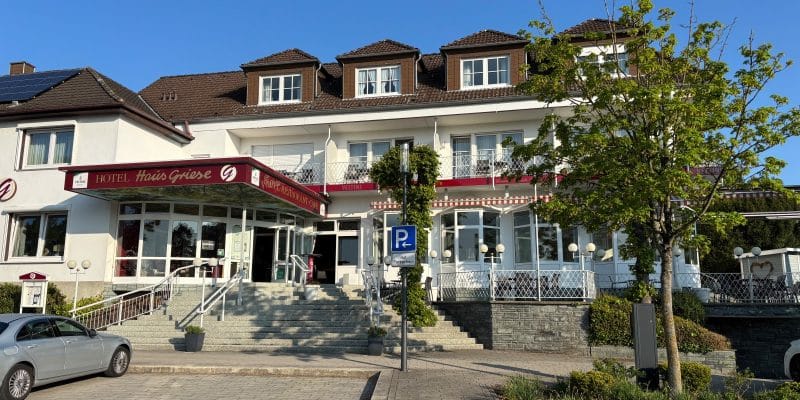 Das Hotel Haus Griese am Möhnesee präsentiert sich als weißes, mehrstöckiges Gebäude mit einer breiten Treppe zum Eingangsbereich und einer Außenterrasse unter strahlend blauem Himmel.