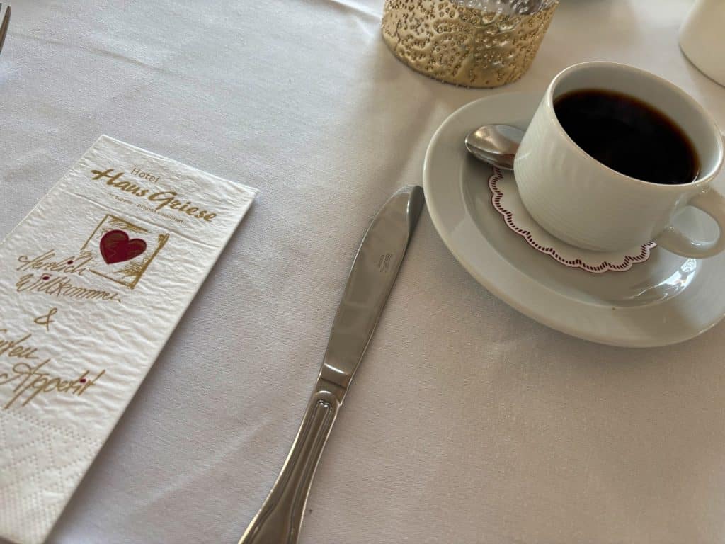 Ein gedeckter Frühstückstisch im Hotel Haus Griese am Möhnesee mit einer Tasse schwarzem Kaffee auf einer Untertasse, einem Messer und einer Serviette mit Herz-Logo.