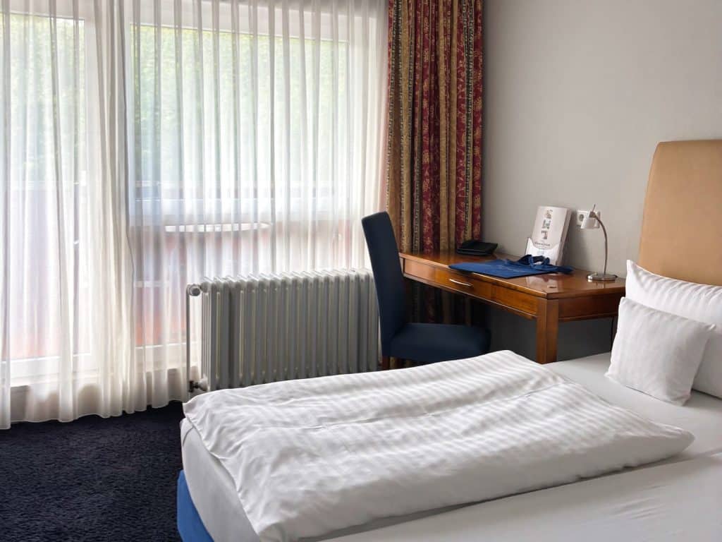 Ein helles Hotelzimmer im Hotel Haus Griese am Möhnesee mit einem gemachten Doppelbett im Vordergrund, einem hölzernen Schreibtisch mit blauem Stuhl an der Wand und einem großen Fenster mit weißen Gardinen.