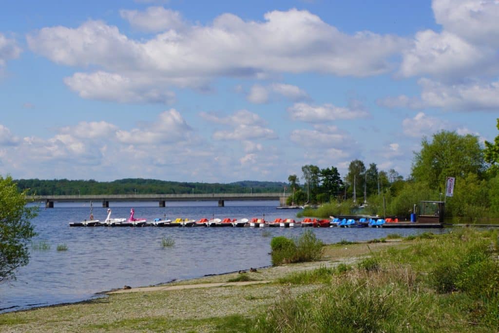 Eine Reihe bunter Tretboote liegt an einem Steg am Ufer des Möhnesees, während im Hintergrund eine lange Brücke das ruhige Wasser unter einem leicht bewölkten blauen Himmel überspannt.