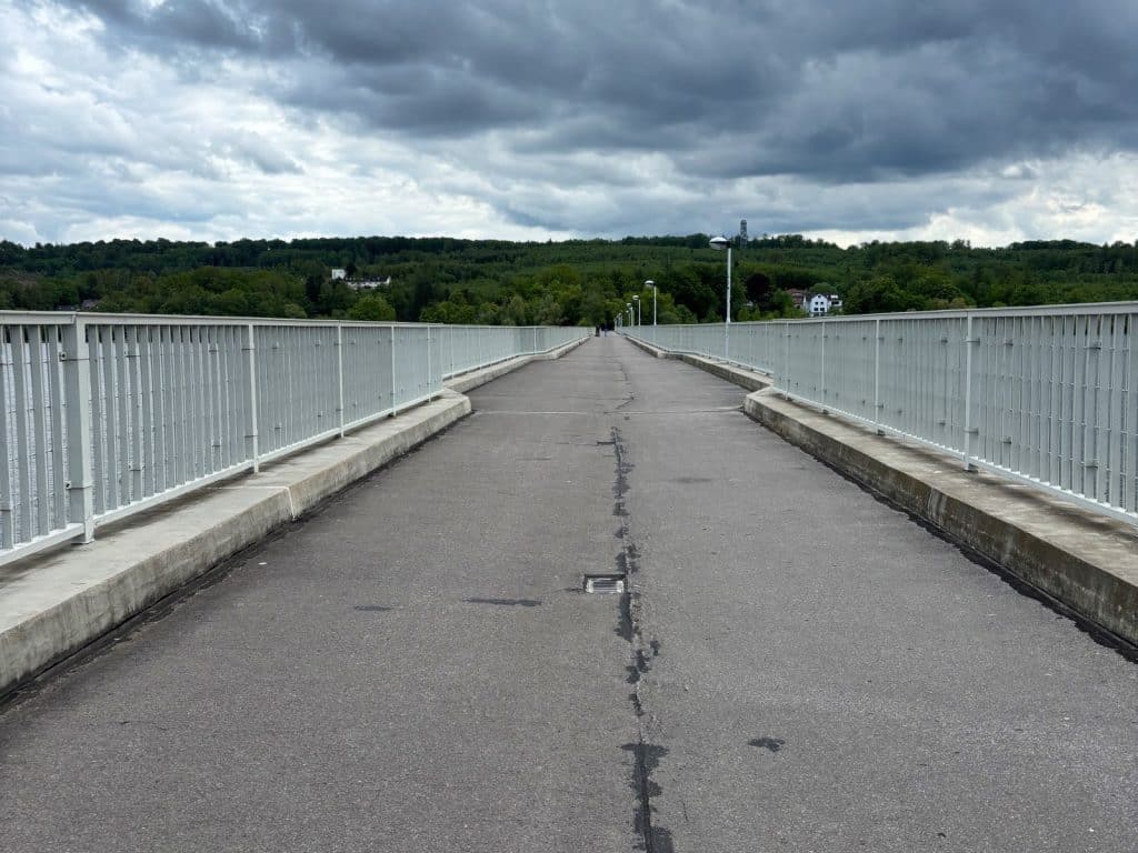 Blick über die asphaltierte Fahrbahn der Möhnesee-Staumauer, die von hellgrauen Metallgeländern flankiert wird und auf bewaldete Hügel unter einem bewölkten Himmel zuführt.