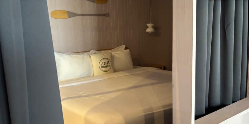 Ein gemütliches Bett in einer Wohnung in Québec mit weißen Kissen, einer dekorativen Kissenrolle mit der Aufschrift „The Lofts Dorchester“ und zwei an der Wand hängenden Holzpaddeln.