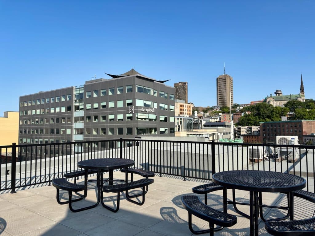Blick von einer Dachterrasse in Québec auf schwarze Metall-Picknicktische vor einem modernen Bürogebäude und der städtischen Skyline unter blauem Himmel.