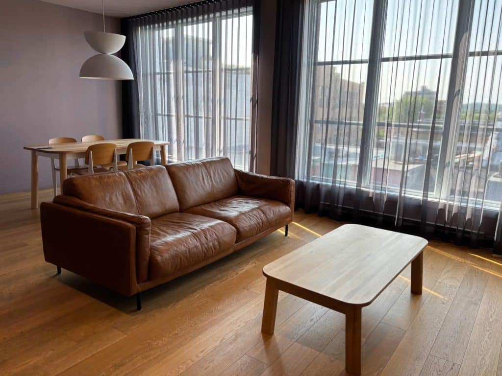 Der Wohnbereich einer Wohnung in Québec ist mit einem braunen Ledersofa, einem hölzernen Couchtisch und einem Essbereich vor großen, bodentiefen Fenstern mit transparenten Vorhängen eingerichtet.