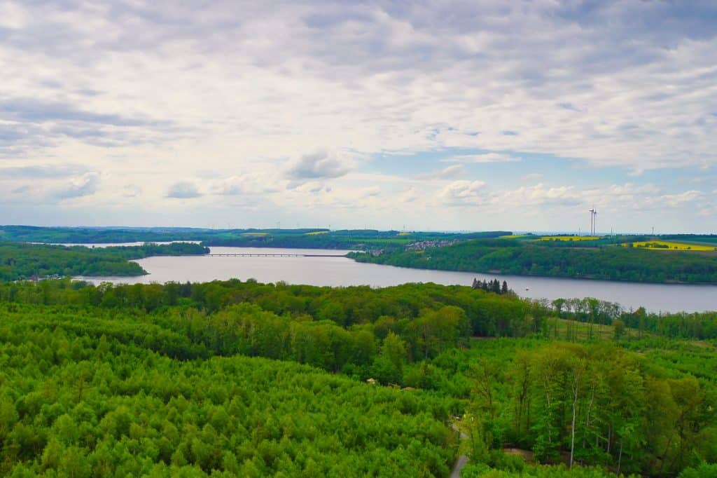 Weite Panoramaaufnahme der hügeligen Waldlandschaft im Sauerland mit Blick auf einen großen Stausee unter einem bewölkten Himmel. Im Hintergrund sind sanfte grüne Hügel, kleine Ortschaften und vereinzelte Windkraftanlagen zu erkennen.