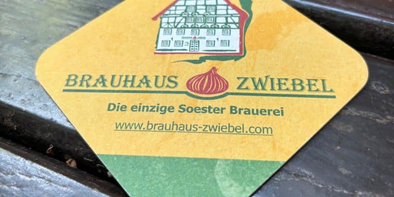 Ein quadratischer Bierdeckel der Brauerei „Brauhaus Zwiebel“ aus Soest liegt auf einem dunklen Holztisch und zeigt eine Illustration eines Fachwerkhauses sowie eine rote Zwiebel. Unter dem Logo stehen der Schriftzug „Die einzige Soester Brauerei“ und die Website-Adresse auf gelbem und grünem Hintergrund.