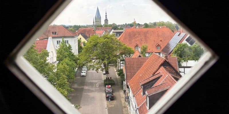 Blick durch ein rautenförmiges Fenster auf eine Straße in Soest mit historischen Fachwerkhäusern, roten Ziegeldächern und den Kirchtürmen von St. Patrokli und St. Petri im Hintergrund.