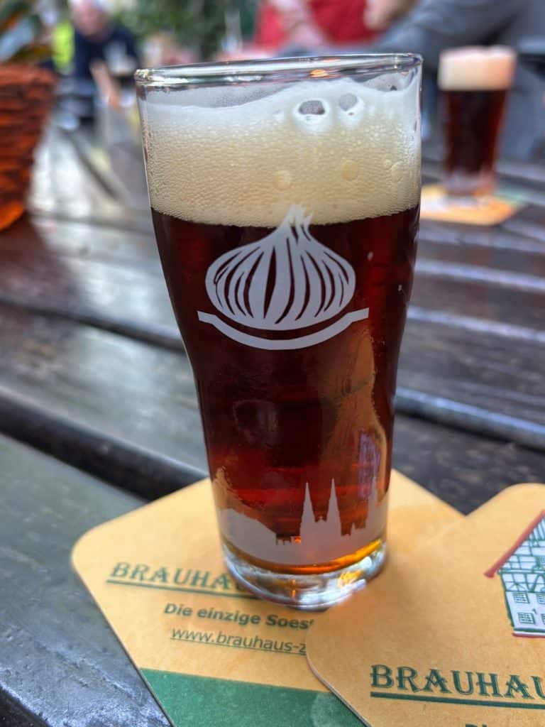 Menüsafari in Soest: Ein Glas dunkles Bier mit einer ausgeprägten Schaumkrone steht auf einem Bierdeckel im Brauhaus Zwiebel in Soest, verziert mit einem weißen Zwiebel-Logo und der Soester Stadtsilhouette.