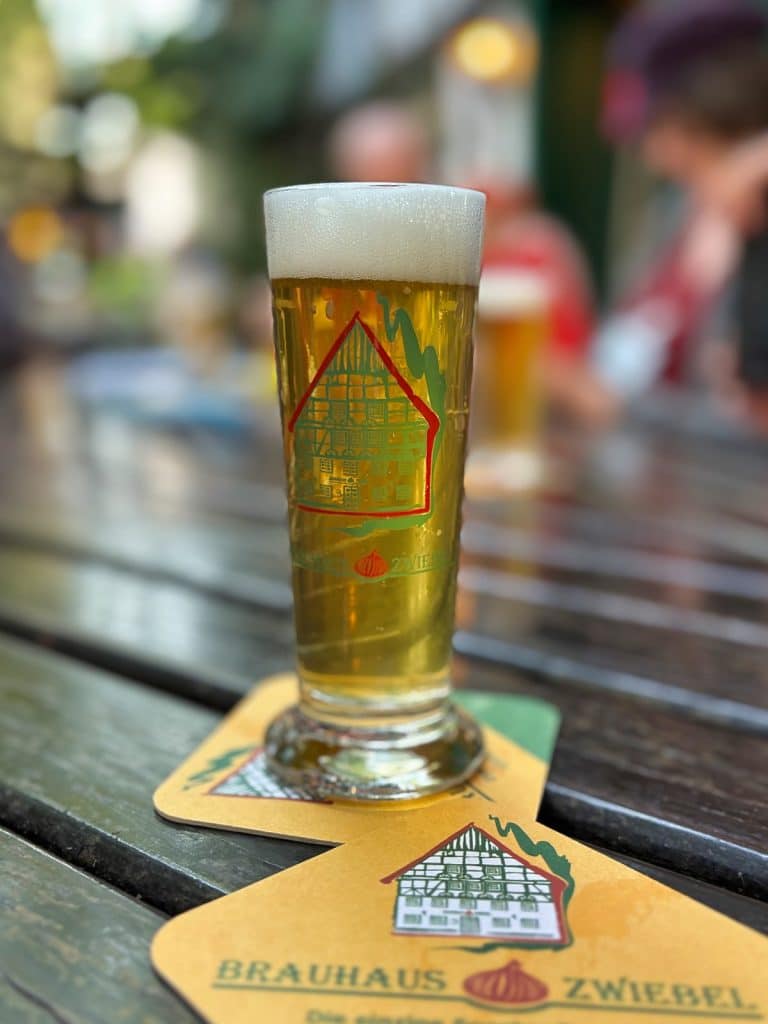 Ein frisch gezapftes Glas helles Bier steht auf einem Bierdeckel des Brauhaus Zwiebel in Soest auf einem rustikalen Holztisch im Freien. Sowohl das Glas als auch der Bierdeckel zeigen das Logo des Brauhauses mit einem Fachwerkhaus und einer roten Zwiebel.
