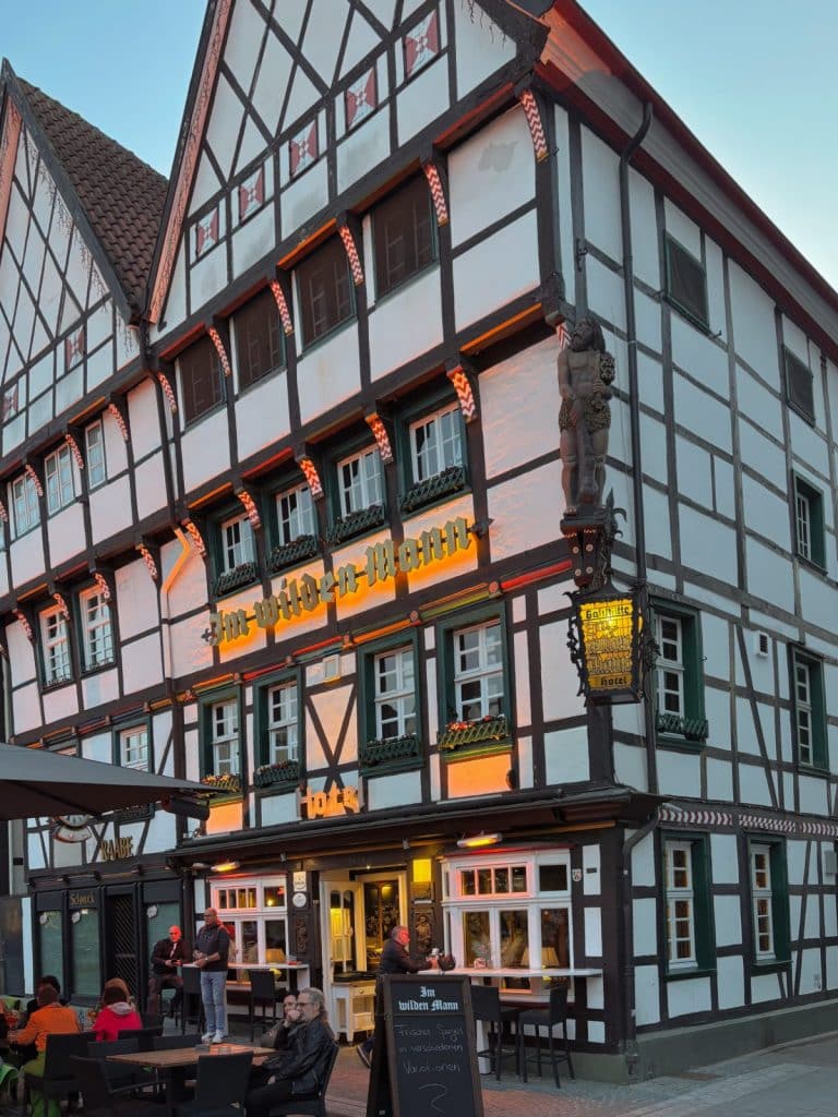Das historische Fachwerkhaus des Hotels und Restaurants „Im Wilden Mann“ in Soest bei Dämmerung, mit beleuchteter Außenwerbung und Gästen, die an Tischen im Außenbereich sitzen.