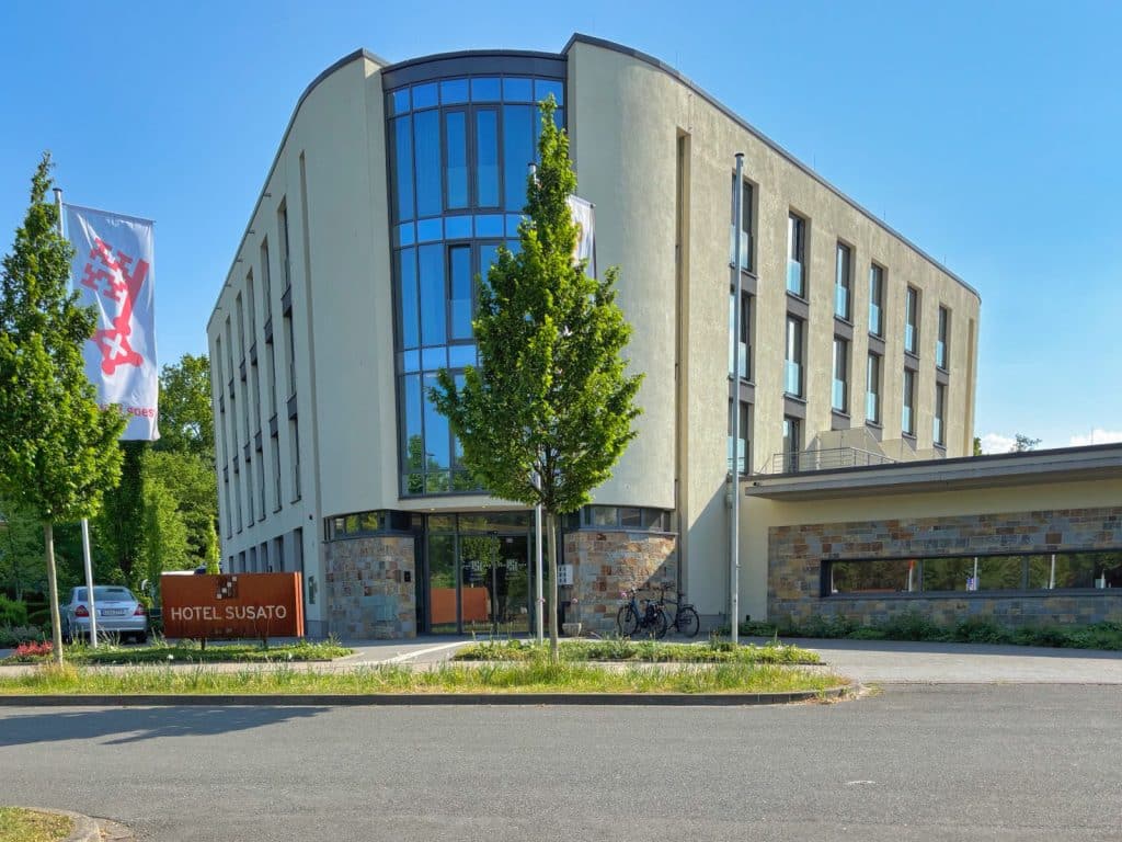 Die Außenansicht des modernen Hotel Susato in Soest zeigt ein helles, mehrstöckiges Gebäude mit einer markanten Glasfront, einem Steinsockel und einer davor gehissten Flagge der Stadt.