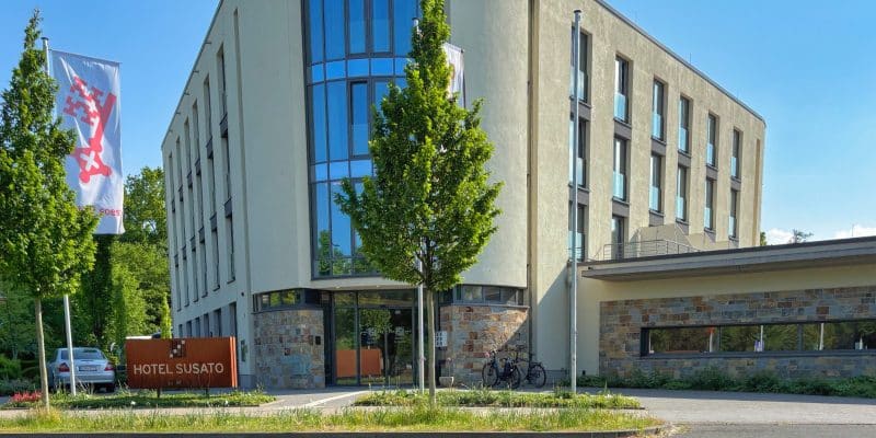 Die Außenansicht des modernen Hotel Susato in Soest zeigt ein helles, mehrstöckiges Gebäude mit einer markanten Glasfront, einem Steinsockel und einer davor gehissten Flagge der Stadt.