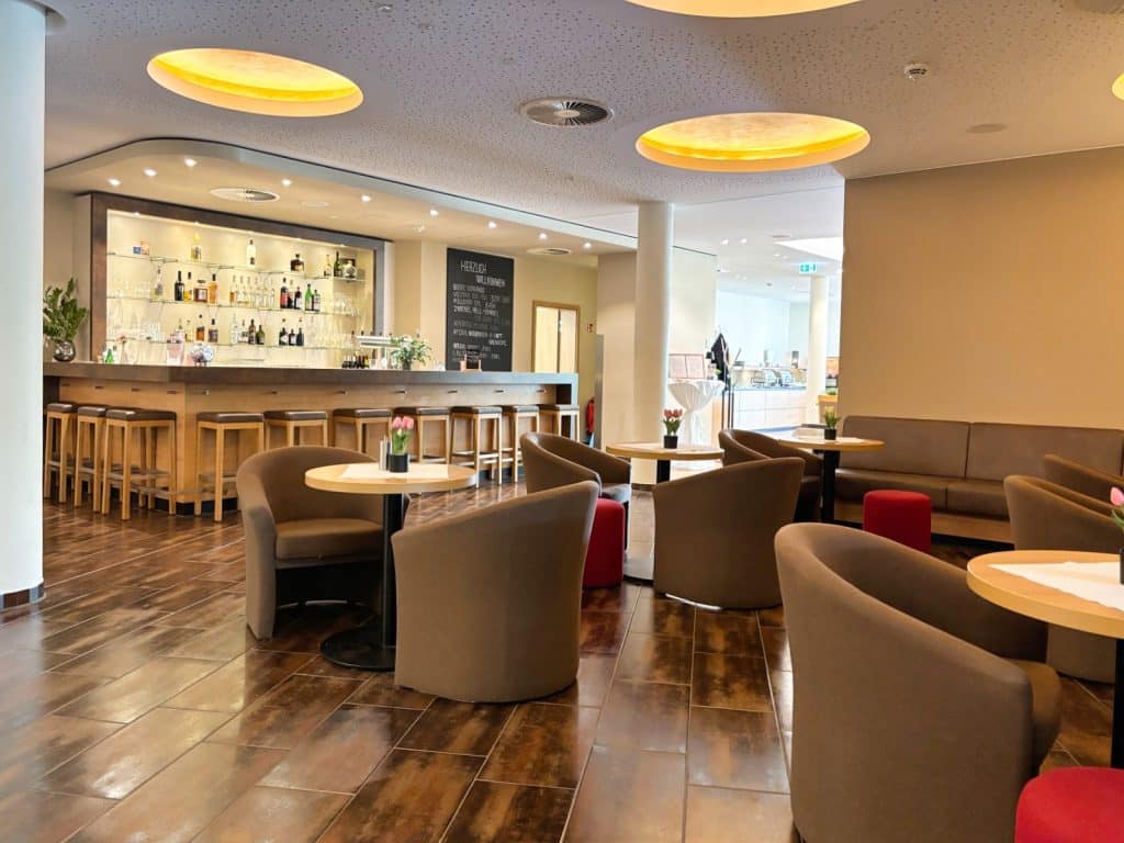 Die moderne Bar des Susato Hotels lädt mit einer beleuchteten Theke, gemütlichen braunen Sesseln an runden Holztischen und warmen, kreisförmigen Deckenleuchten zum Verweilen ein.